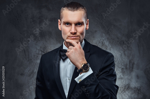 Elegantly dressed handsome man wearing a black classical suit and bow tie, touching his face and looking at a camera.