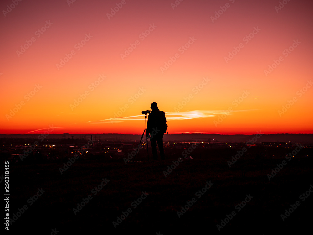 Image of a silhouette of a man with tripot and camera standing on a hill during colorful sunset
