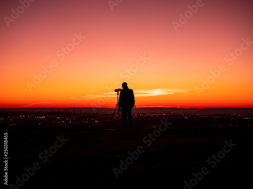 Image of a silhouette of a man with tripot and camera standing on a hill during colorful sunset