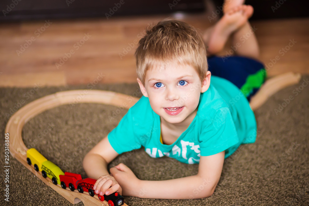 Fototapeta premium happy baby, blonde with blue eyes, a little boy of five years in the mint (green / blue) t-shirt in shorts barefoot near the couch in the apartment igret fun with wooden railway