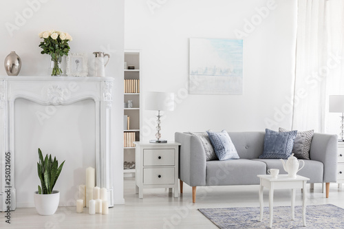 White table on carpet in front of grey sofa in elegant flat interior with plants and poster. Real photo