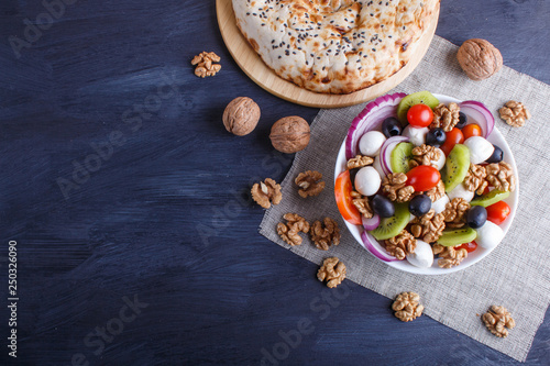 Salad with cherry tomatoes, mozzarella cheese, black olives, kiwi, and walnuts on black wooden background.