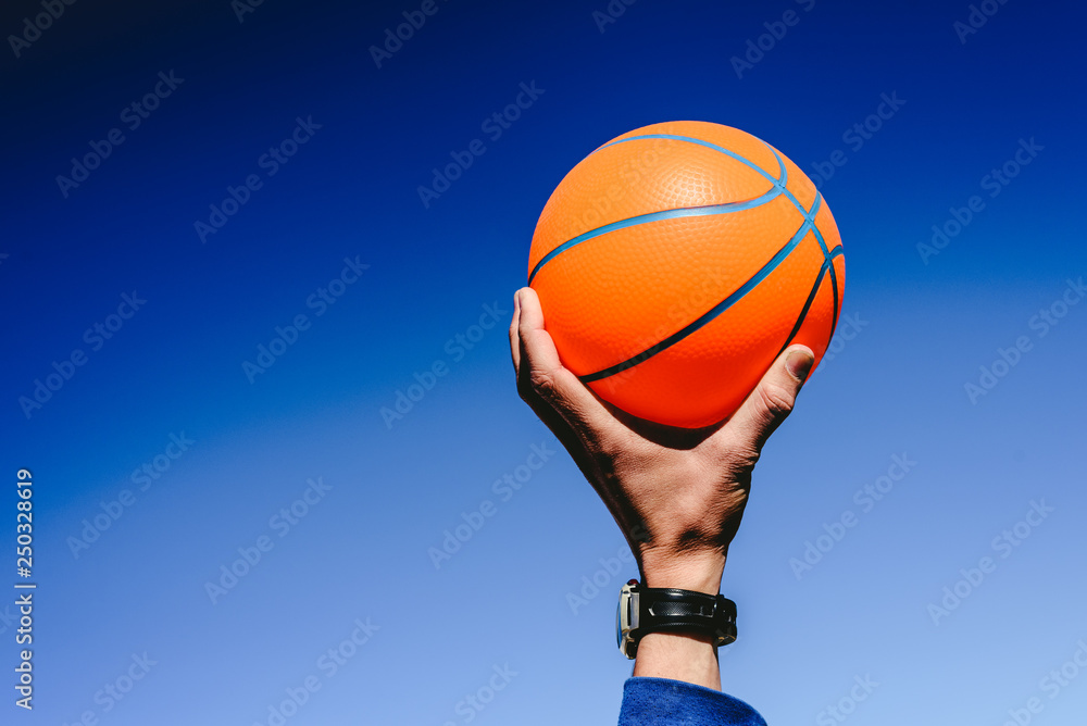 Hand holding an orange basketball ball on blue sky background, invitation to play, copy space free area.