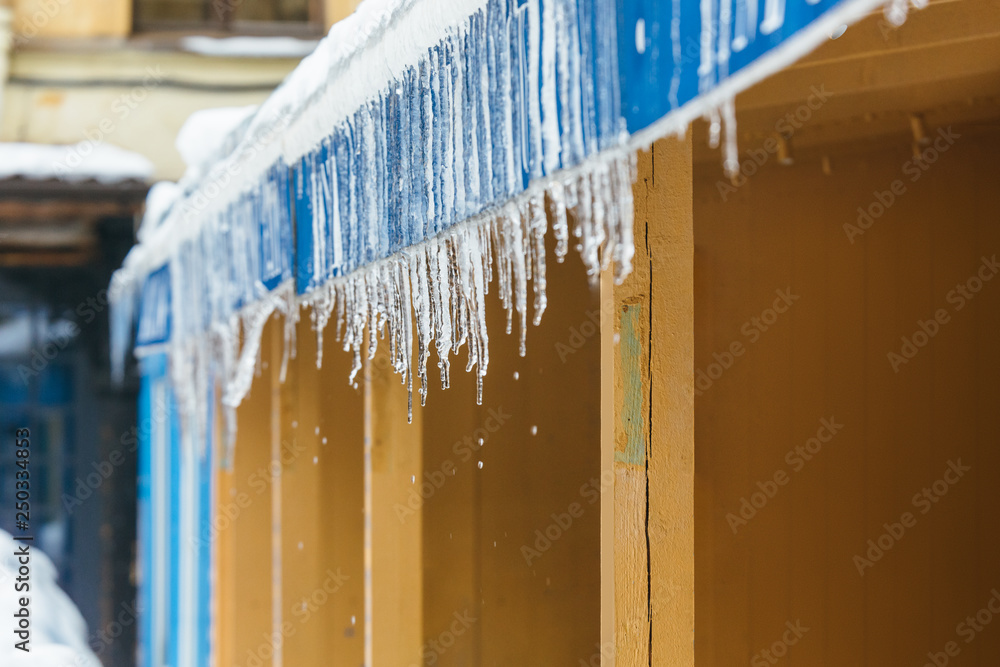 Icicles hanging from roof of the building, drops are dripping from them/ danger for humans, sudden warming, poor thermal insulation, ice stalactite, formation of icicles/ winter weather concept. Icicles hanging from roof of the building, drops are dripping from them/ danger for humans, sudden warming, poor thermal insulation, ice stalactite, formation of icicles/ winter weather concept.