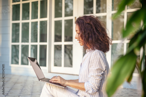 A young beautiful woman is sitting on the terrace with a laptop in the background of her home.