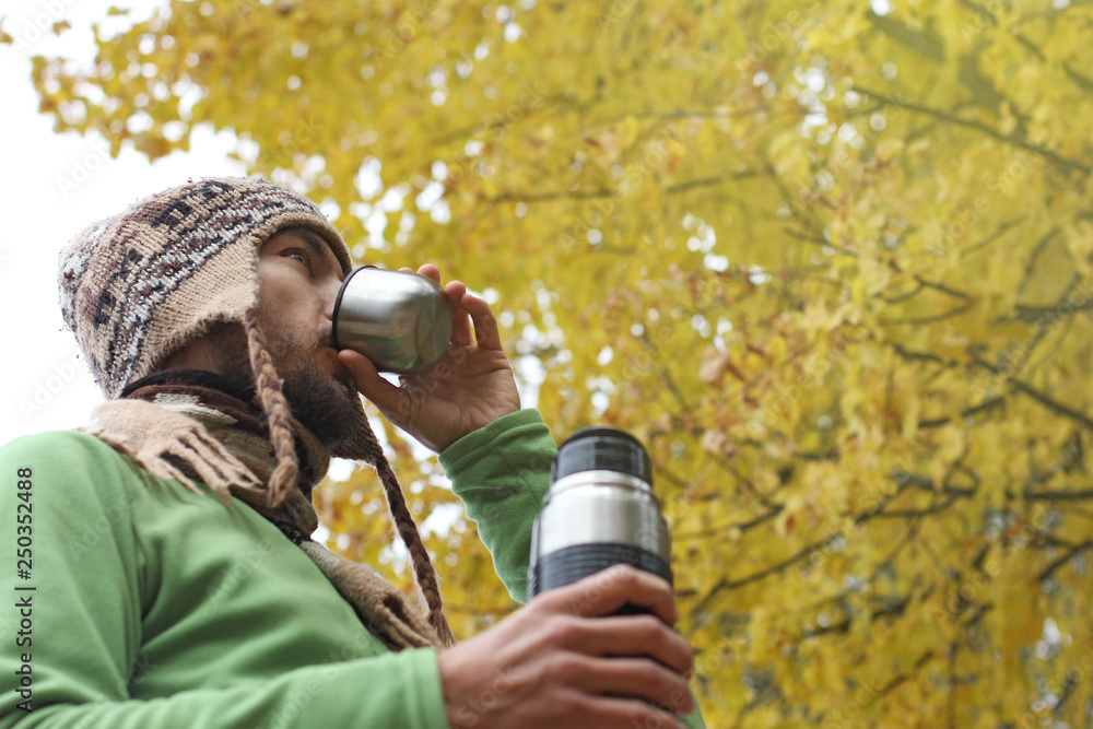 Naklejka premium bearded man in wool knitted hat drinks hot tea or coffee from mug, bottom side view, background of yellow autumn foliage