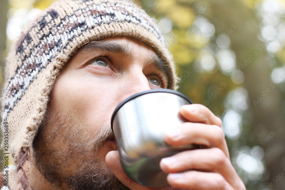bearded man in wool knitted hat intently drinks hot tea or coffee from mug, side view on face, background of forest bearded man in wool knitted hat intently drinks hot tea or coffee from mug, side view on face, background of forest