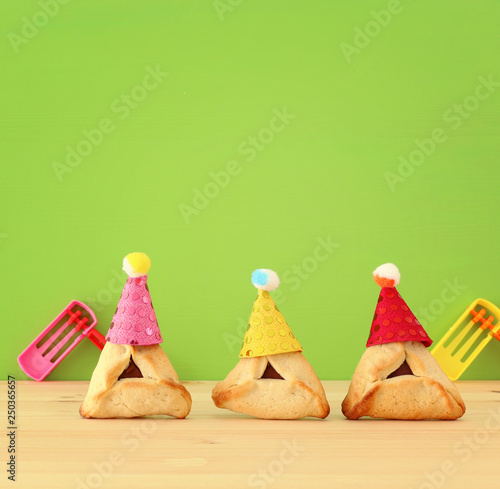 Purim celebration concept (jewish carnival holiday). Traditional hamantaschen cookies with cute clown hats and noisemaker over wooden table.