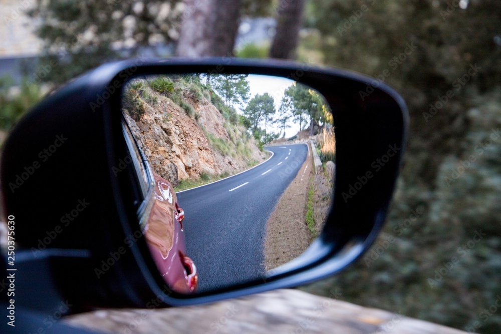 Car driving travel concept - car rear view mirror view of a mountain road with rocks and trees Car driving travel concept - car rear view mirror view of a mountain road with rocks and trees