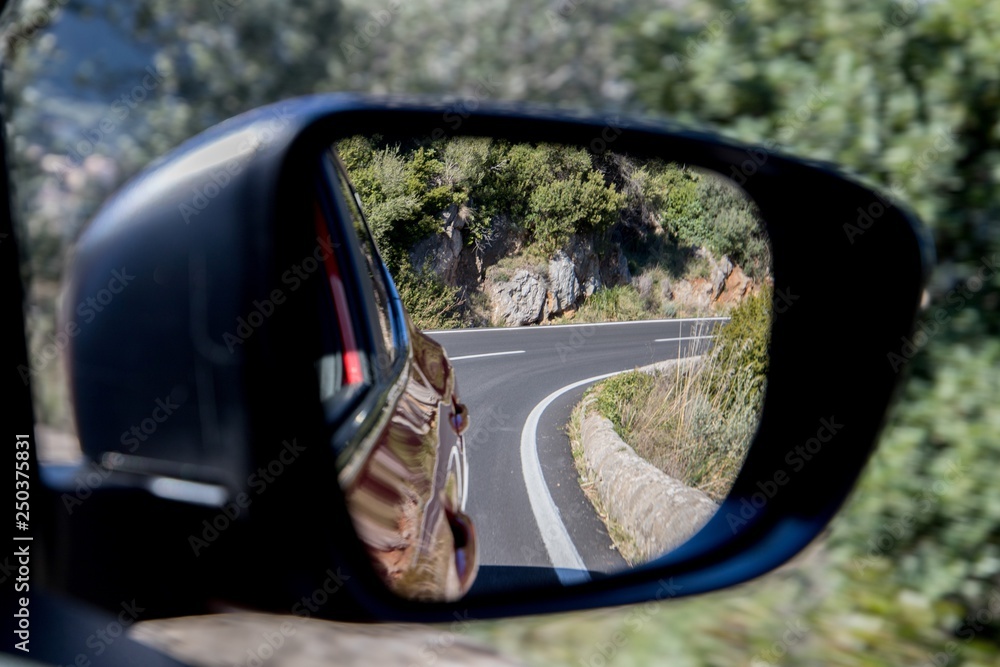 Car driving travel concept - car rear view mirror view of a mountain road with rocks and trees Car driving travel concept - car rear view mirror view of a mountain road with rocks and trees