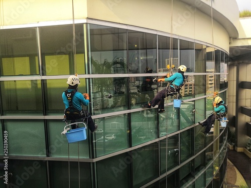 Rope access cleaning worker wearing safety harness hard hat working at height descending on rope performing washes a hospital complex glass wall. 