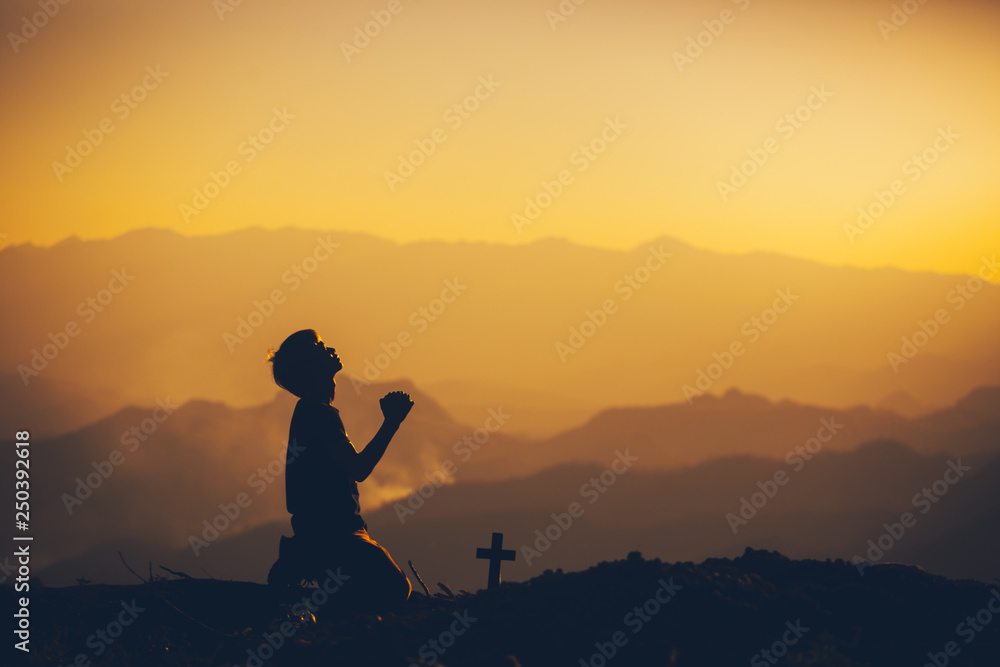 Young man kneeling down and praying with christian cross at sunset background. christian silhouette concept. - obrazy, fototapety, plakaty 