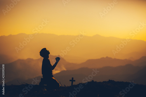 Murais de parede Young man kneeling down and praying with christian cross at sunset background