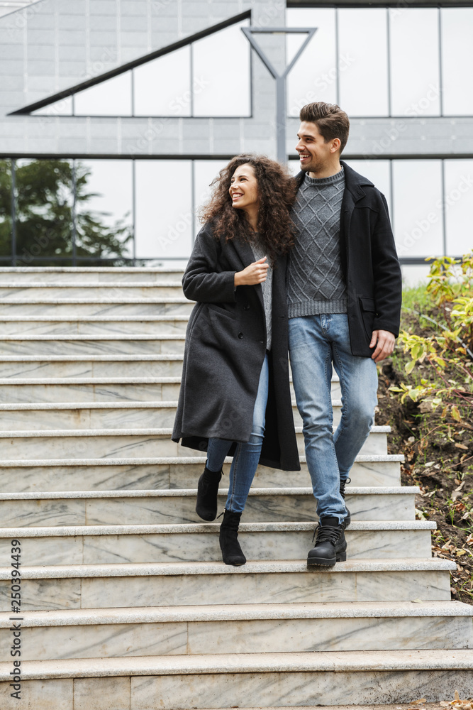 Full length photo of caucasian couple man and woman 20s in warm clothes, walking down gray stairs outdoor
