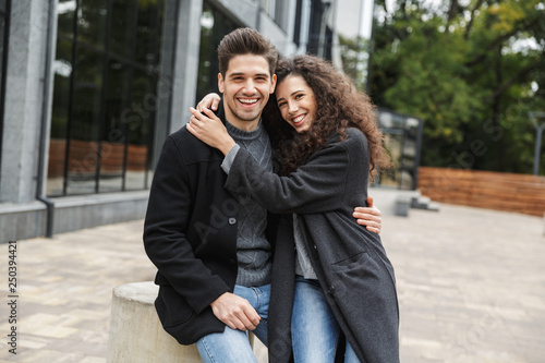Photo of happy couple man and woman 20s hugging while, standing over gray building outdoor