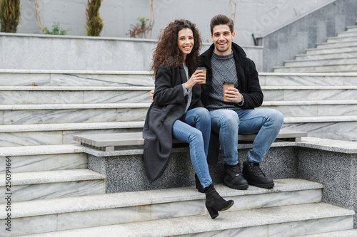 Image of young couple man and woman 20s in warm clothes, drinking takeaway coffee while sitting on bench outdoor