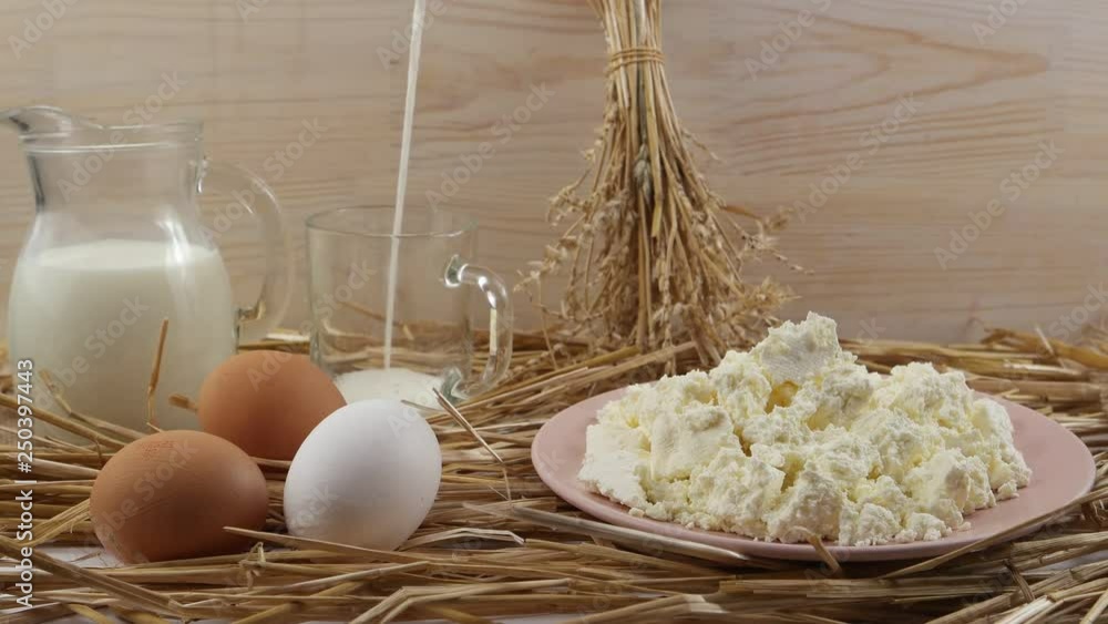 Milk filling a glass, glass jug of milk, chicken eggs and a plate with cottage cheese on a background of straw and wooden board Milk filling a glass, glass jug of milk, chicken eggs and a plate with cottage cheese on a background of straw and wooden board