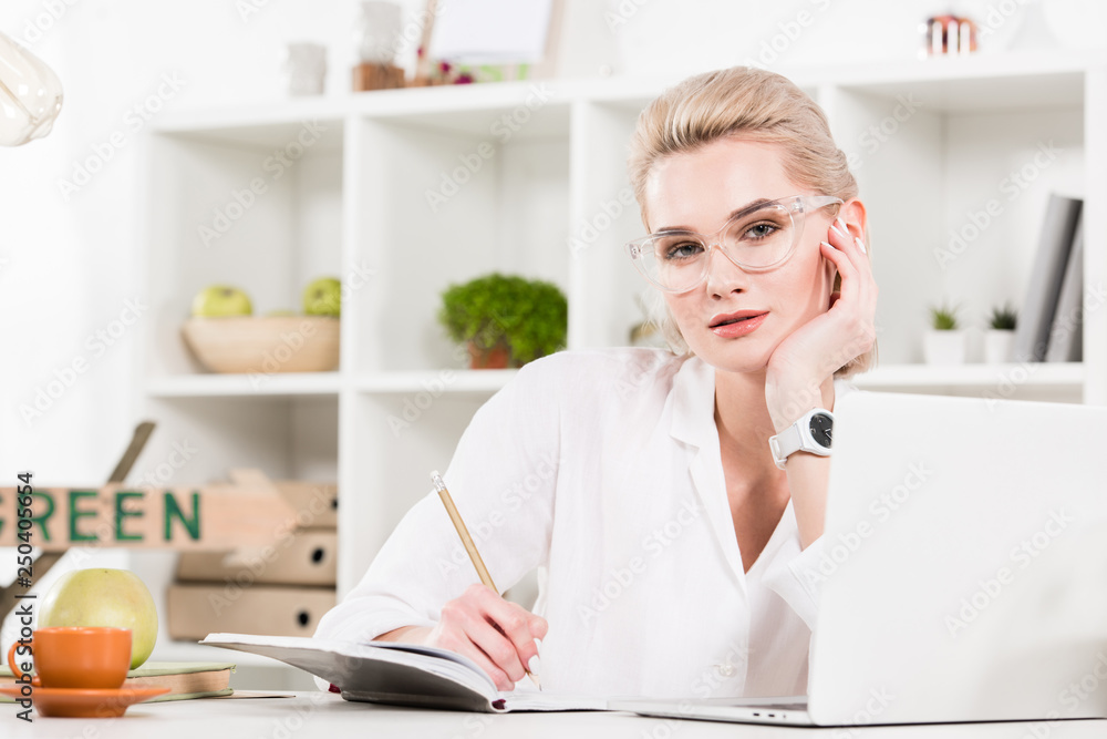 woman in glasses writing in notebook near laptop in office, environmental saving concept woman in glasses writing in notebook near laptop in office, environmental saving concept