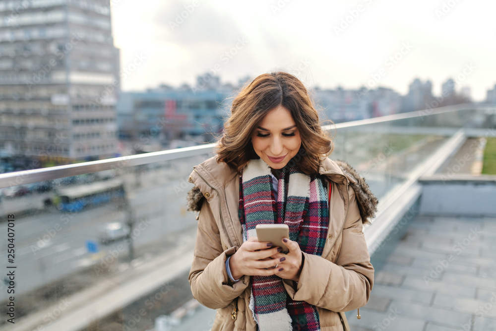 Portrait of beautiful brunette in jacket and with scarf standing on rooftop and using smart phone. Portrait of beautiful brunette in jacket and with scarf standing on rooftop and using smart phone.