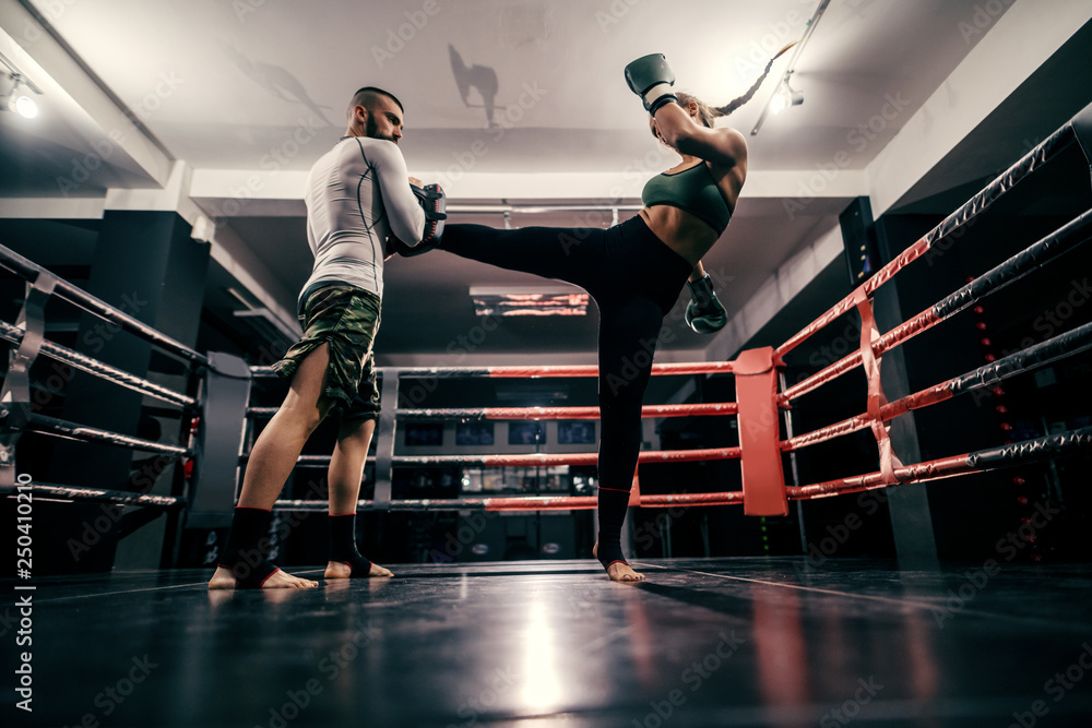 Φωτογραφία Powerful caucasian boxer girl in sportswear, with bandage on feet and boxing gloves on hands practicing with trainer in ring