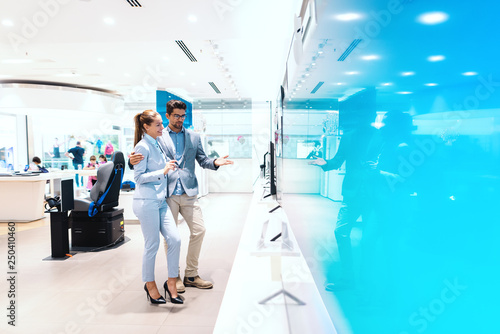 Beautiful multicultural couple dressed elegant standing and looking for new tv. Tech store interior.