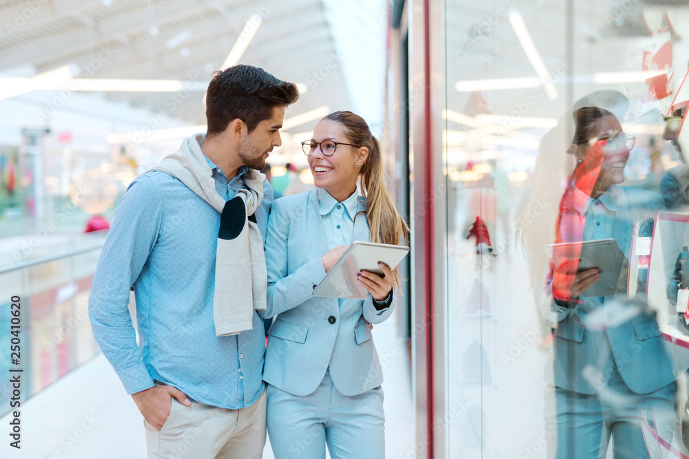 Happy couple standing in front of shop window and looking for something to buy. Woman holding tablet. Shopping mall interior. Happy couple standing in front of shop window and looking for something to buy. Woman holding tablet. Shopping mall interior.