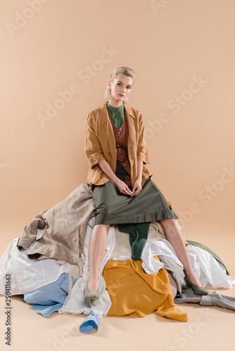 blonde woman sitting on pile of clothing on beige background, environmental saving concept