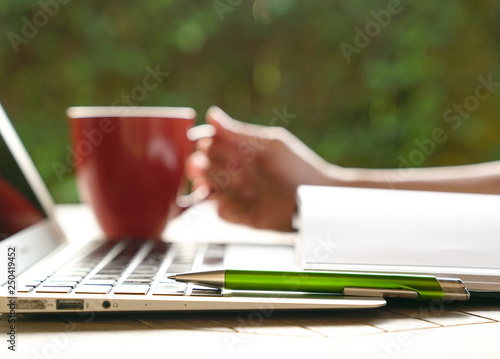 Work concept: hand on red mug with laptop, note pad in view and focus on pen