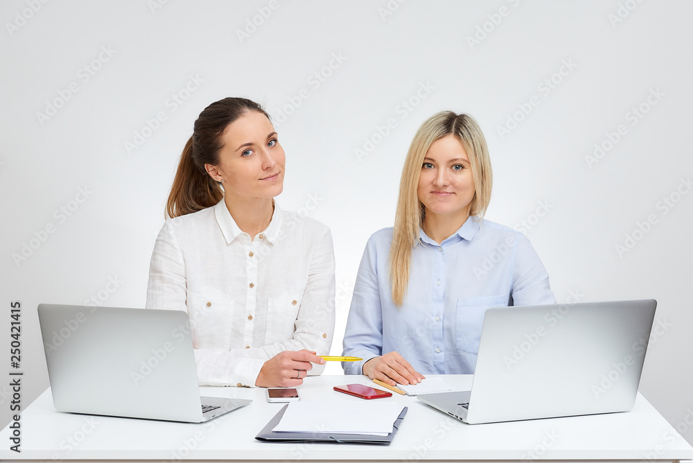 Cute brunette female manager with smile and her blonde caucasian colleague sits by the desk in the office with the white background. Cute brunette female manager with smile and her blonde caucasian colleague sits by the desk in the office with the white background.