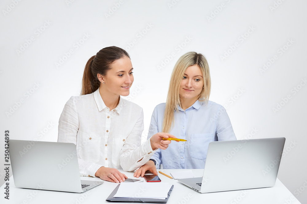 Brunette female manager with a smile and her blonde caucasian colleague works together sits by the desk in the office with the white background. Brunette female manager with a smile and her blonde caucasian colleague works together sits by the desk in the office with the white background.