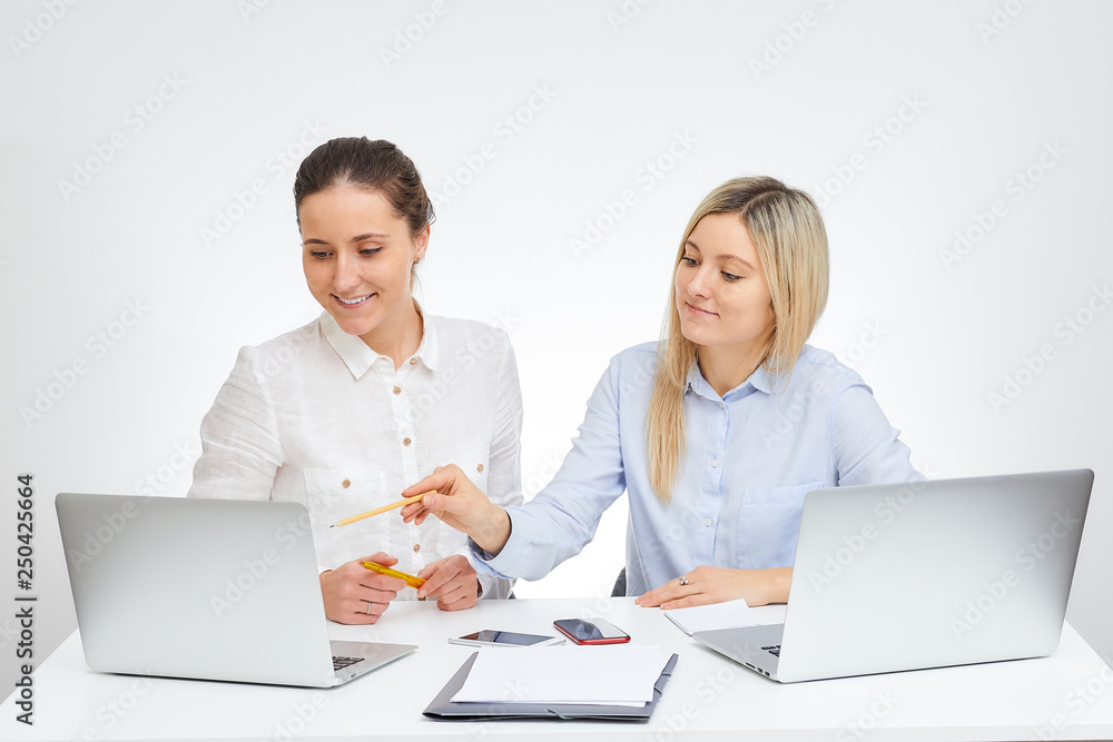 Happy brunette female manager with a smile and her blonde caucasian colleague works together sits by the desk in the office with the white background. Happy brunette female manager with a smile and her blonde caucasian colleague works together sits by the desk in the office with the white background.