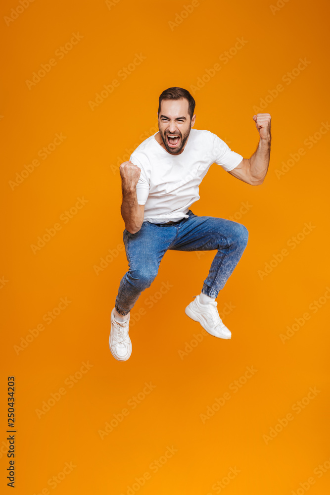 Full length photo of young guy in t-shirt and jeans jumping and having fun, isolated over yellow background