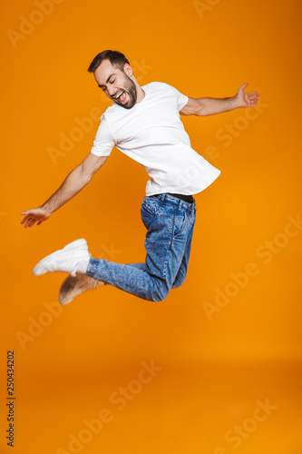 Full length photo of positive guy in t-shirt and jeans jumping and having fun, isolated over yellow background