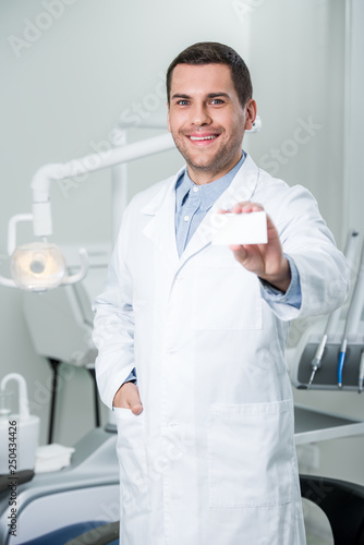 cheerful dentist standing with hand in pocket and holding blank card