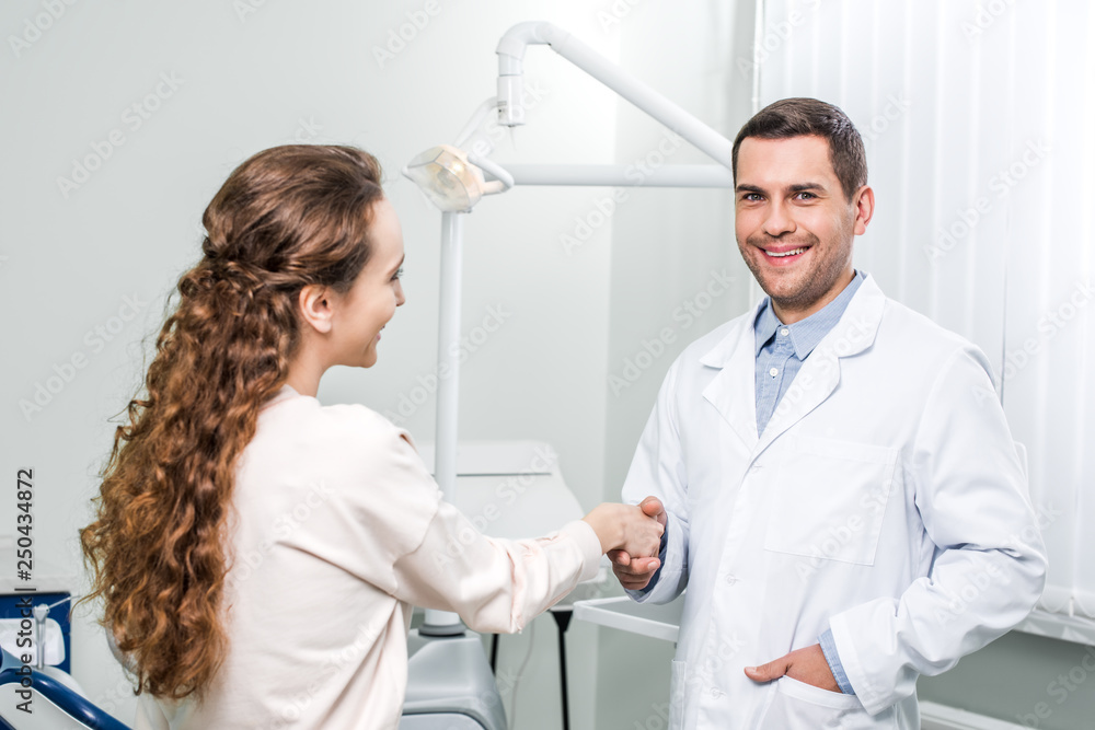 smiling dentist standing with hand in pocket and shaking hands with female patient smiling dentist standing with hand in pocket and shaking hands with female patient