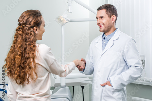 cheerful dentist standing with hand in pocket and shaking hands with female patient