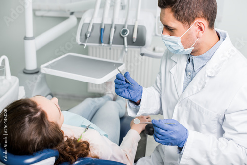 serious dentist in latex gloves and mask holding dental equipment near female patient
