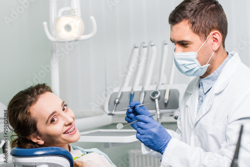 dentist in latex gloves and mask holding dental equipment near beautiful woman with braces