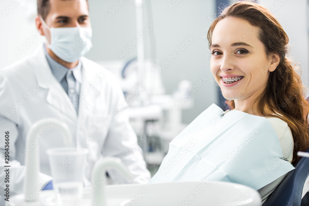 selective focus of woman in braces smiling with dentist standing in mask on background selective focus of woman in braces smiling with dentist standing in mask on background