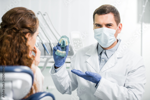 selective focus of dentist in mask and latex gloves showing teeth model near female patient