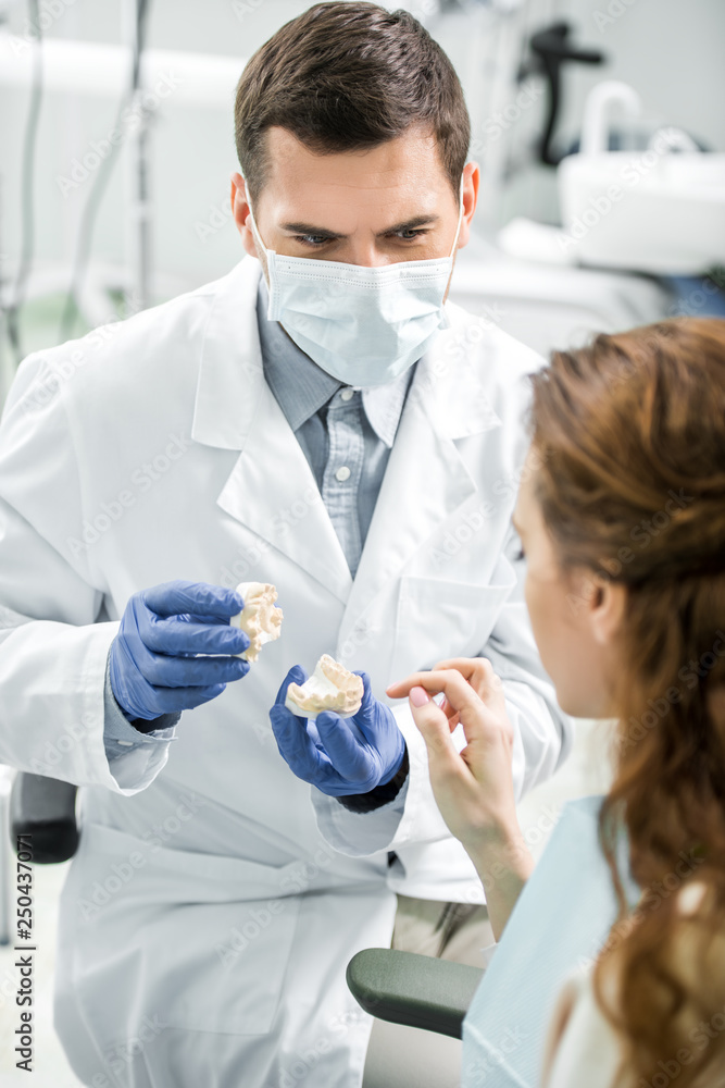 selective focus of dentist in latex gloves and mask holding teeth model near female patient