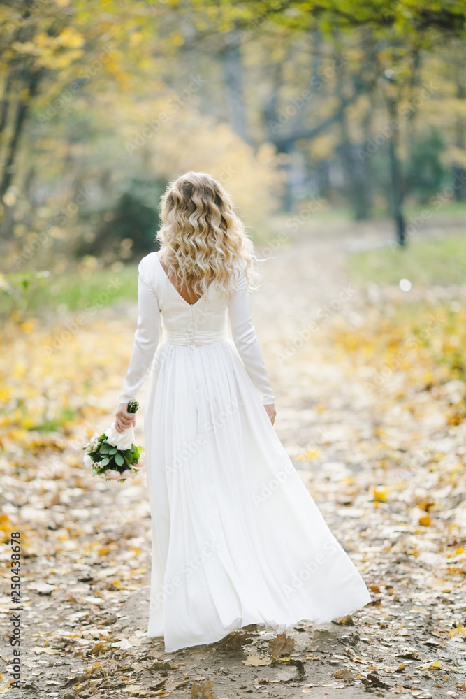 Look from behind at charming bride posing in autumn park covered with fallen leaves