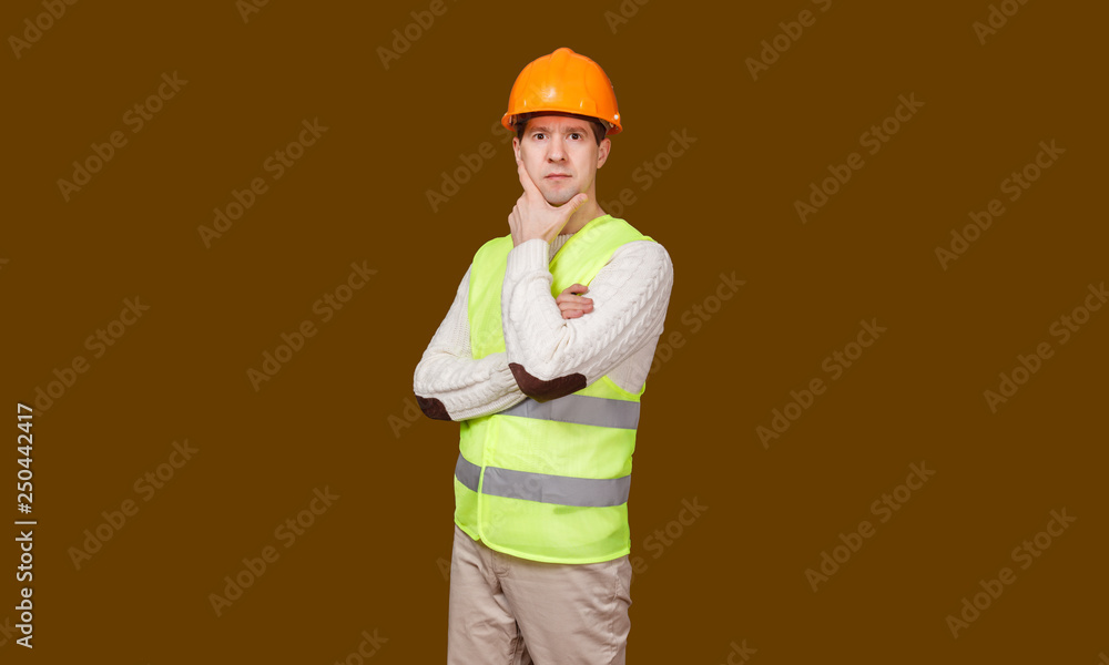 The thoughtful man in a construction helmet and a vest, looks in the camera, the crossed hands at the head