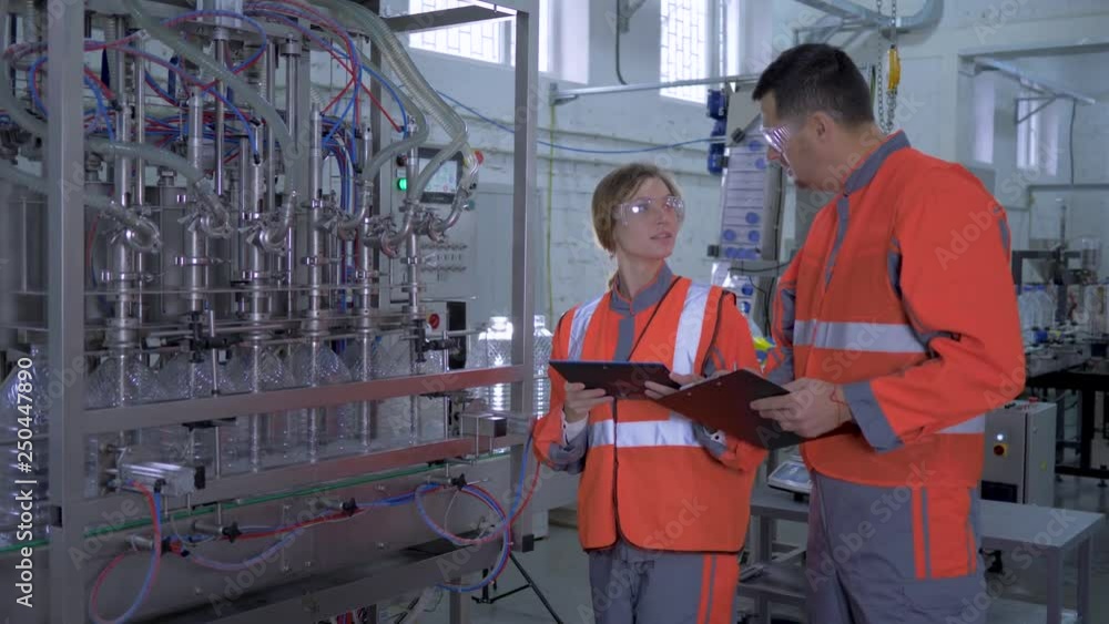 factory profession, young woman and man Industry workers into coveralls with digital tablet in hand near automatic conveyor line with bottling of mineral water in plastic bottles on production plant factory profession, young woman and man Industry workers into coveralls with digital tablet in hand near automatic conveyor line with bottling of mineral water in plastic bottles on production plant