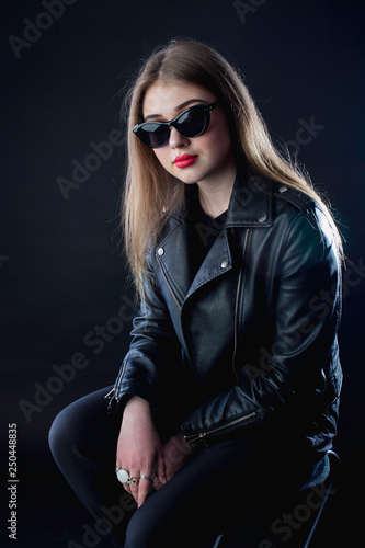 young girl in a black leather jacket and sunglasses on a dark background in the studio sitting