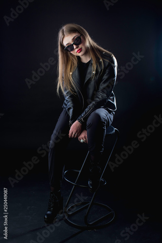 young girl in a black leather jacket and sunglasses on a dark background in the studio sitting