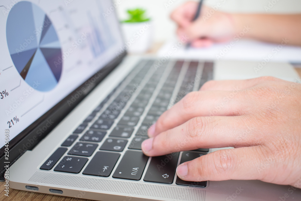 The side view of the hand is working on the laptop and notes the financial graph report in the office. The side view of the hand is working on the laptop and notes the financial graph report in the office.