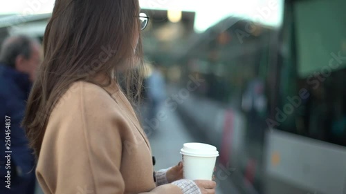 Slow motion profile european attractive female entrepreneur holding take-away morning coffee heading work waiting train station platform, commute home, living lively city, standing relaxed casually photo