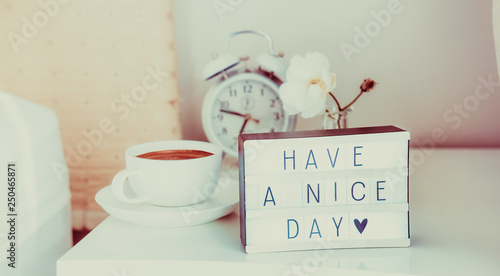 Fotografia, Obraz Have a nice day message on lighted box, alarm clock, cup of coffee and flower on the bedside table in sun light