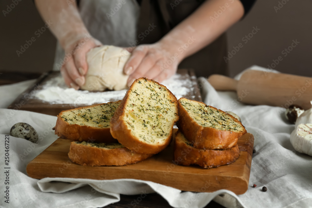 Tasty cut garlic bread on table and woman kneading dough in kitchen